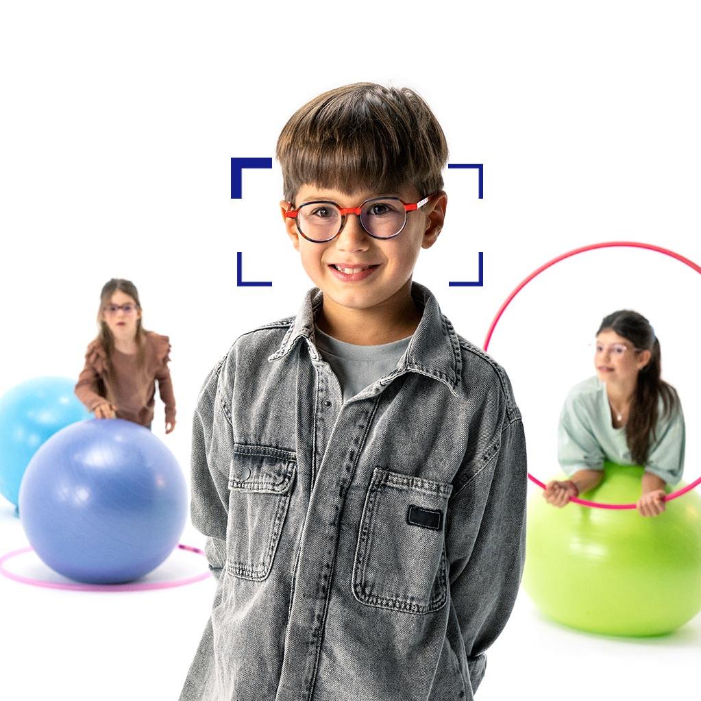 Un garçon aux cheveux marrons avec des lunettes rondes qui porte des verres ZEISS MyoCare est debout au premier plan et sourit pour la photo. À l’arrière-plan, deux filles portant des verres ZEISS MyoCare jouent avec des cerceaux et des ballons de gymnastique.