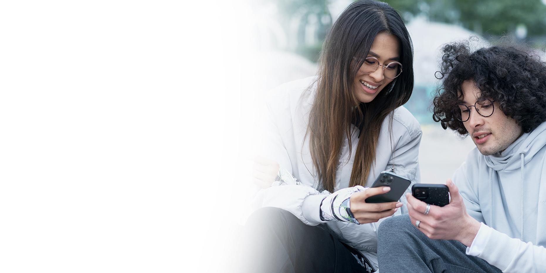 Zwei junge Leute, eine lächelnde Frau mit langen braunen Haaren und ein Junge mit lockigen schwarzen Haaren und ZEISS SmartLife Brille, schauen auf ihre Handys. 