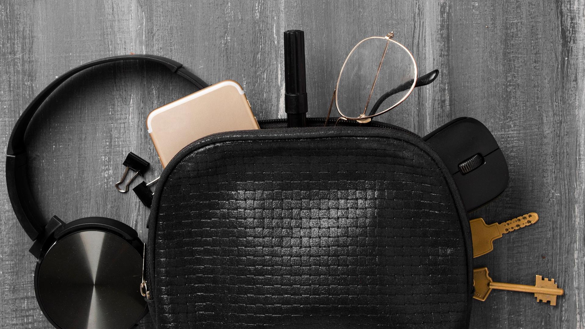 Eine kleine schwarze Tasche, aus der Kopfhörer, ein Handy, Schlüssel, Büroklammern, ein Stift, eine PC-Maus und eine Brille mit ZEISS Brillengläsern mit DuraVision Beschichtung herausgerutscht sind und nun teilweise auf dem grauen Boden liegen.