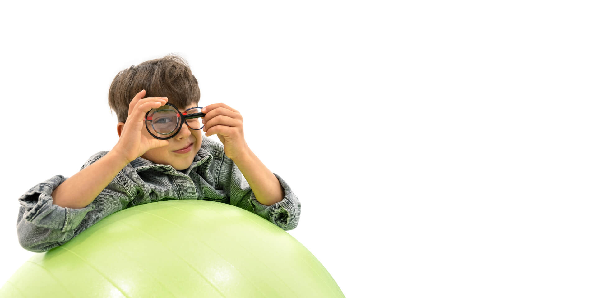 Ein Junge mit ZEISS MyoCare Brillengläsern lehnt sich an einen Gymnastikball und hält sich eine Lupe vor ein Auge.