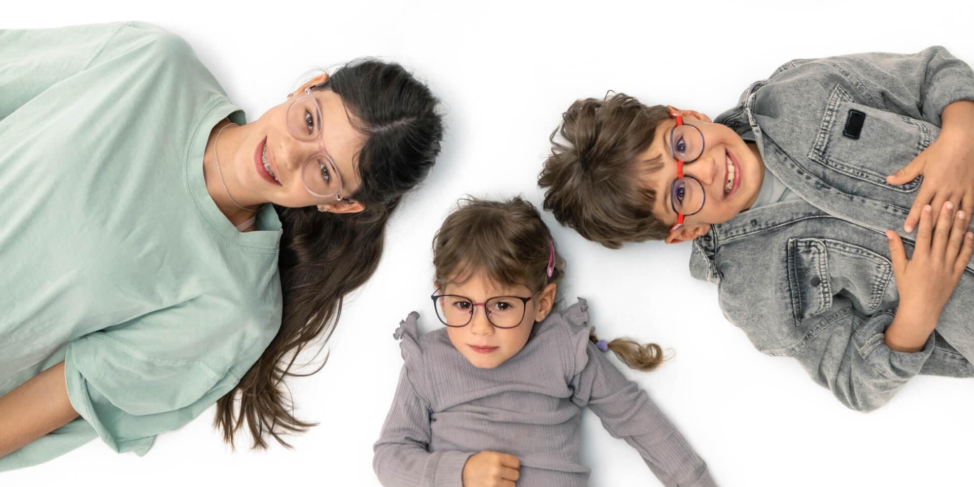 Ein Teenager und zwei kleinere Kinder liegen auf dem Boden und tragen Brillen mit ZEISS Brillengläsern für die Behandlung von Kurzsichtigkeit.