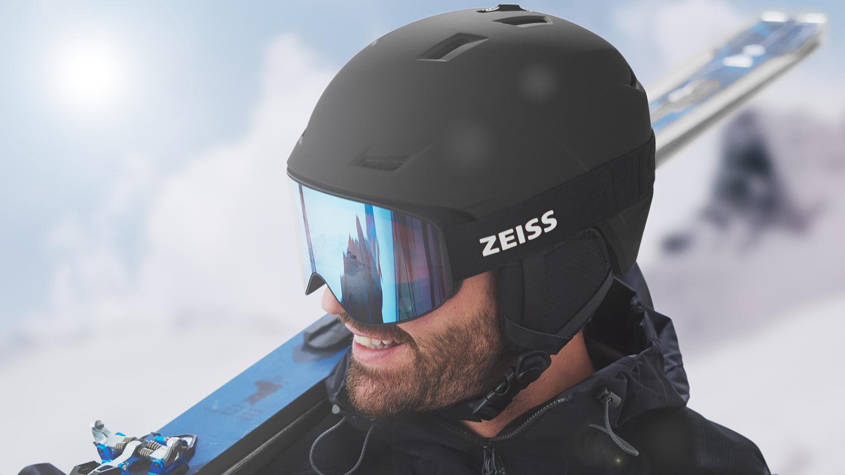 Un homme porte un casque noir et un masque de ski ZEISS bleu effet miroir, et des skis sur son épaule droite.