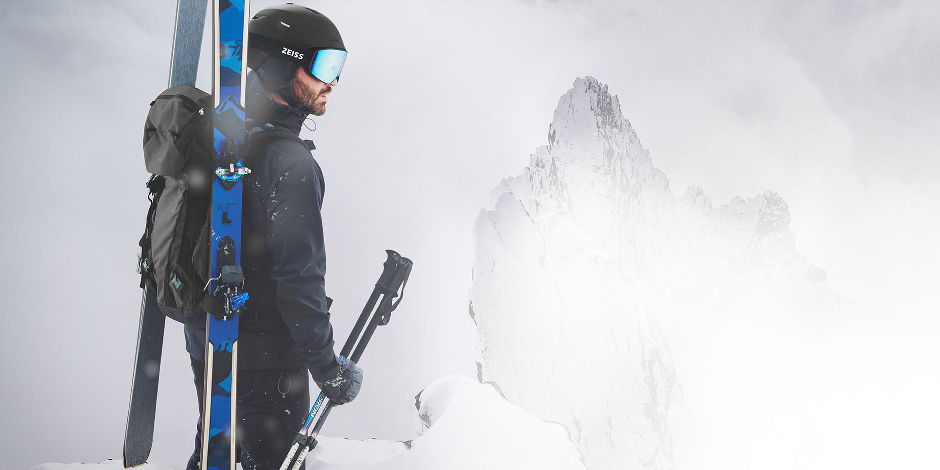 Un homme porte un casque noir et un masque de ski ZEISS à verre bleu miroir, des skis et un sac à dos sur son dos et tient ses bâtons de ski dans sa main droite. Il se tient debout au sommet d’une montagne.