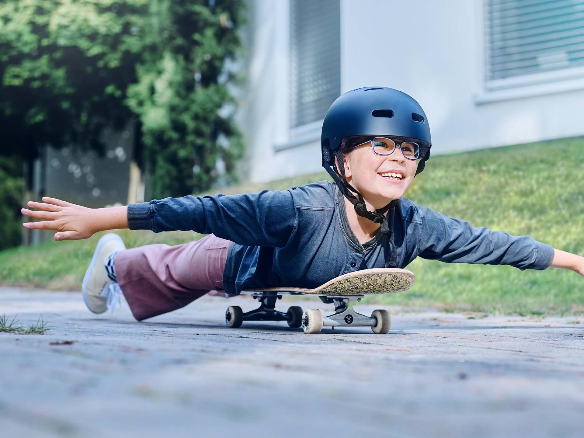 Une fillette portant un casque et des lunettes, allongée sur une planche de skate, descend une rue, bras tendus.