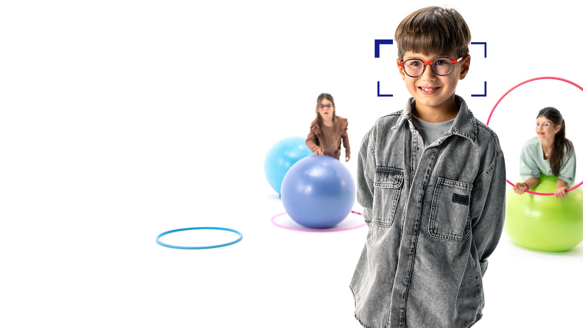 Ein Junge mit braunen Haaren, runder Brille und ZEISS MyoCare Brillengläsern steht im Vordergrund und lächelt in die Kamera. Im Hintergrund sind zwei Mädchen mit ZEISS MyoCare Brillengläsern zu sehen, die mit Reifen und Gymnastikbällen spielen.